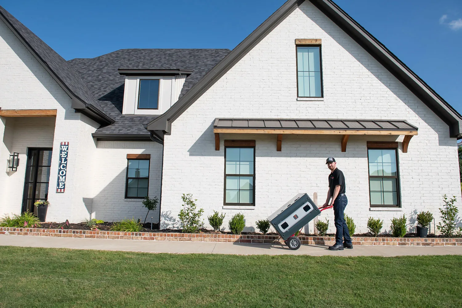 Man moving large appliance on hand truck in front of modern white brick house with lawn