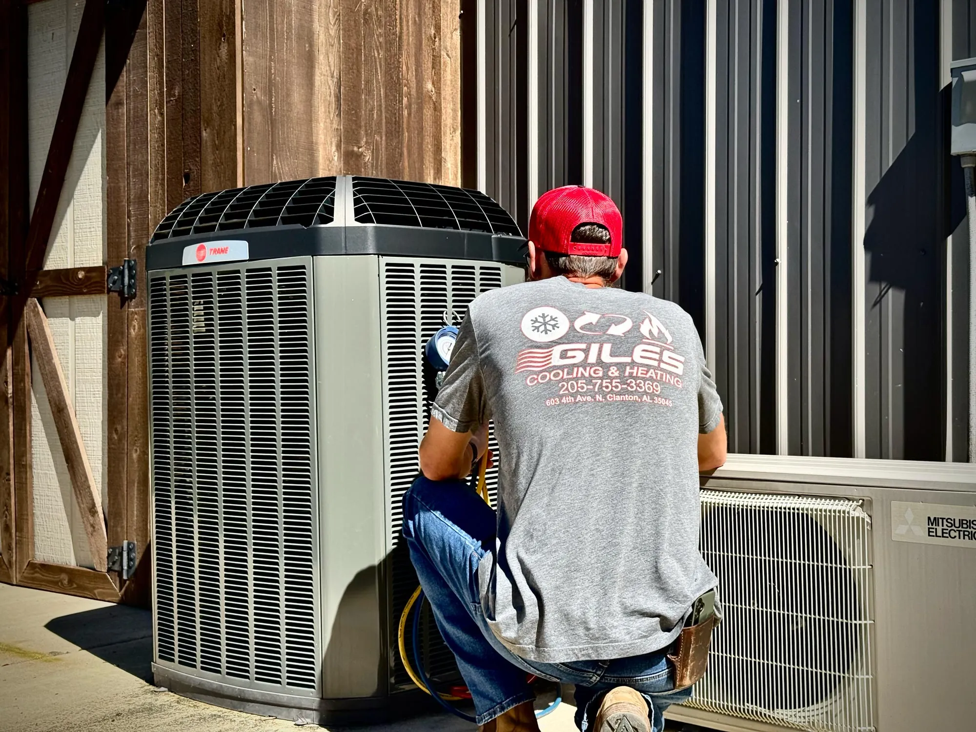 A man in a red cap kneeling to service a Trane outdoor HVAC unit beside a wooden and metal building