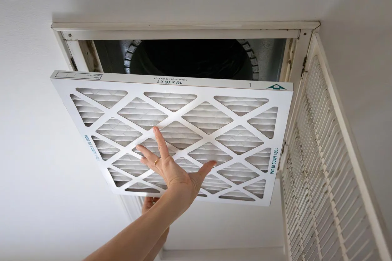 Person replacing HVAC air filter in ceiling return vent, hands installing a 16x16 pleated filter.